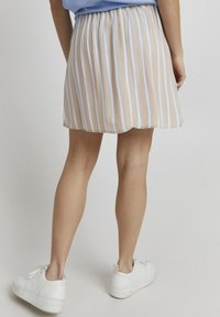 Gonna plissettata beige chiaro con strisce verticali blu chiaro e bianche. L'orlo ha un bordo curvo. Abbinata a sneakers bianche.
