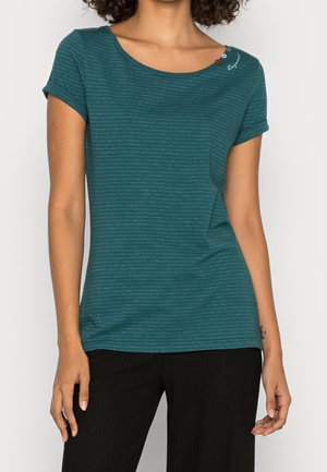 Frau trägt ein türkisfarbenes T-Shirt mit kurzen Ärmeln mit dünnen weißen horizontalen Streifen und schwarzen Hosen, steht vor einem schlichten Hintergrund.