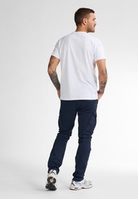 Vit kortärmad t-shirt, marinblå cargobyxor med sidofickor och grå sneakers. Modellen står i profil mot en enkel bakgrund.