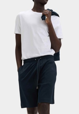 Persoon draagt een wit T-shirt met korte mouwen, marineblauwe short met trekkoord, houdt een marineblauwe jas over de schouder tegen een effen achtergrond.
