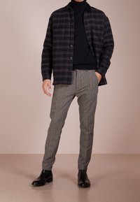Chemise à carreaux noirs et rouges sur un col roulé noir, associée à un pantalon à carreaux gris et des bottes chelsea noires. Textures lisses et coupe ajustée.