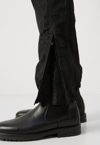 Schwarze Hosen mit einem schimmernden, strukturierten Finish, die über Reißverschlüsse an den Unterschenkeln verfügen. Kombiniert mit schwarzem Leder-Schuhwerk.