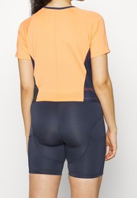Maglietta sportiva arancione a maniche corte con tessuto testurizzato, abbinata a pantaloni corti attillati blu scuro con dettagli delle cuciture e tasche laterali.