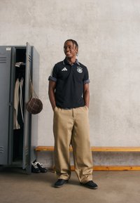 Mladý muž ve sportovním oblečení se usmívá v šatně vedle otevřené skříňky s oblečením a zavěšeným fotbalovým míčem.