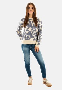 Pull en tricot crème avec un motif abstrait bleu, col rond et poignets côtelés, associé à un jean bleu et des baskets beige.