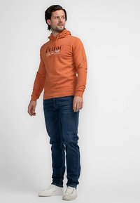 Felpa arancione con testo stampato, cappuccio con coulisse e polsini a costine. Abbinata a jeans blu scuro e sneakers bianche. Sfondo semplice.