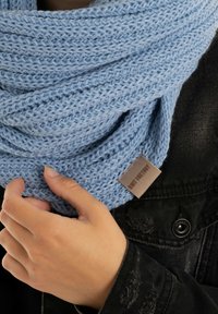 Lichtblauwe gebreide sjaal met een gestructureerde weef, vastgehouden in een hand, met een klein bruin etiket dat aan de rand is bevestigd. Zwarte jas met zichtbare stiksels.
