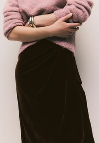 Rosa, flauschiger Pullover über einem dunklen Samtrock mit drapierten Details. Goldene Metallarmreifen schmücken das Handgelenk. Weiche Texturen und satte Farben.