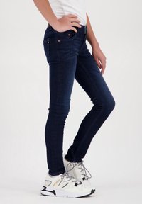 Dunkelblaue Skinny-Jeans aus Denim, mit Fronttaschen und dezenten Hardware-Details. Tragen mit weißen Sneakers und einem taillierten Shirt.
