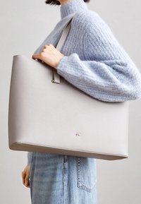 Bolso tote de color gris claro, hecho de cuero texturizado, con una forma plana y una correa larga. Presenta detalles en metal dorado.