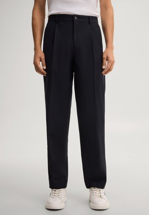Mann trägt schwarze, maßgeschneiderte Hose mit vorderen Falten und weißen Sneakers, steht vor einem einfarbigen hellen Hintergrund.