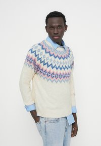 Πουλόβερ σε κρεμ απόχρωση με ροζ και μπλε γεωμετρικά σχέδια, που φοριέται πάνω από ένα ανοιχτό μπλε πουκάμισο με ρίγες. Μανσέτες και γιακάς ριμπ. Άνετη εφαρμογή.