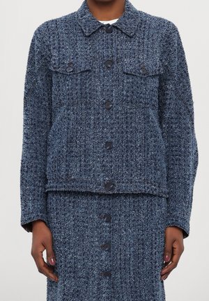 Mujer que lleva una chaqueta azul texturizada con botones y bolsillos de solapa en el pecho, y falda a juego, de pie con las manos relajadas a los lados.