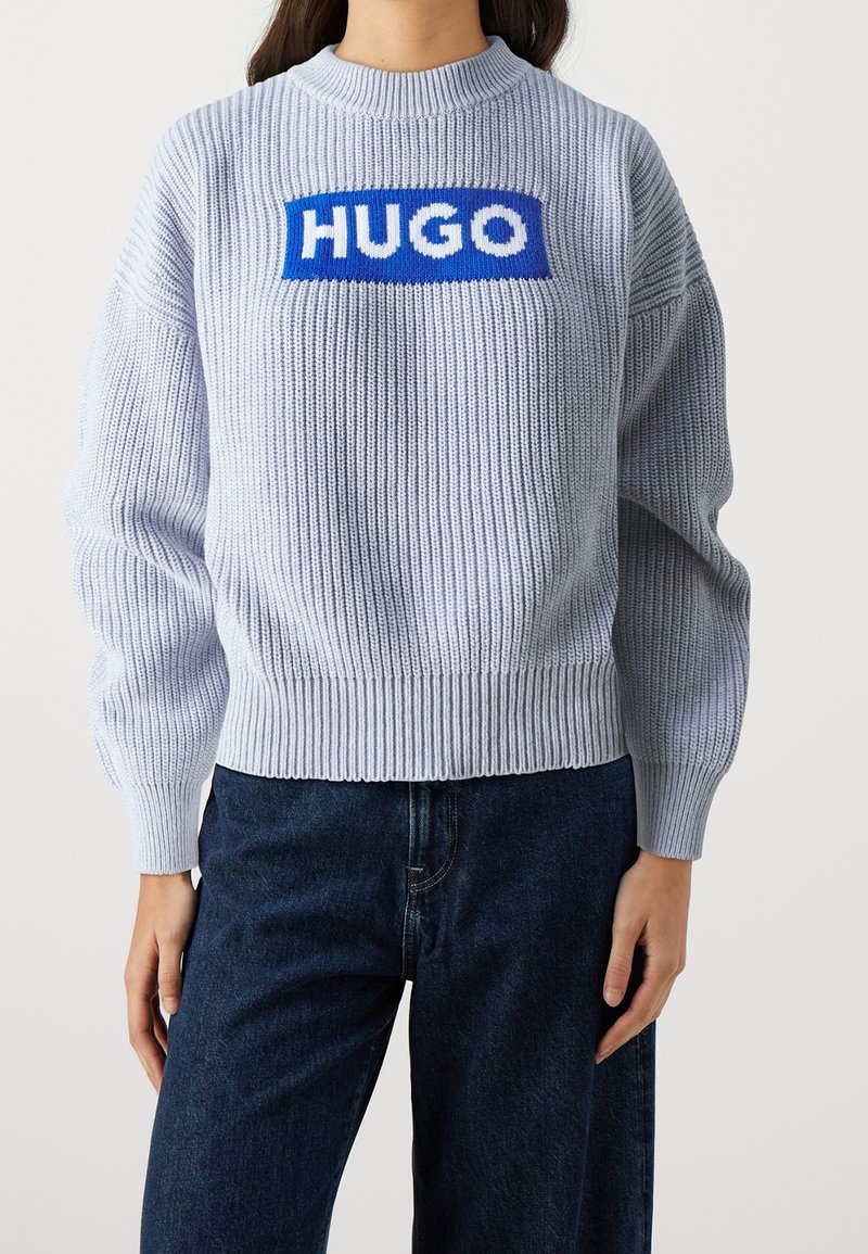 Lichtgrijze gebreide trui met ribgebreide manchetten en onderkant, voorzien van een gedurfde blauwe en witte "HUGO" logo over de borst. Ontspannen pasvorm.