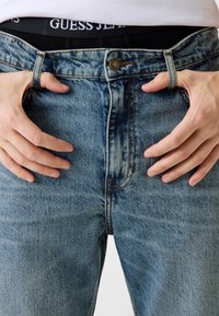 Niebieskie jeansy z denimu z wyblakłym wykończeniem, fasonem z pięcioma kieszeniami i rozporkiem na guziki. Pasek na talii ozdobiony metką "GUESS JEANS".