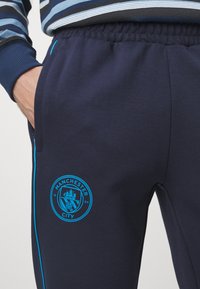 Tumši zili sporta bikses, izgatavotas no mīkstas auduma, ar Manchester City logo gaiši zilā krāsā un kontrastējošām sānu svītrām. Iekļauts elastīgs jostasvīts.