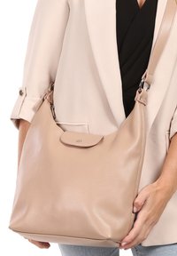 Sac à main en cuir beige avec une finition texturée, une forme arrondie, une bandoulière réglable et un rabat de fermeture avec un petit logo.