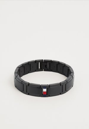 Pulseira de link em metal preto com segmentos retangulares e um pequeno detalhe de bandeira nas cores vermelho, branco e azul em um dos segmentos.