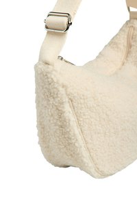 Bolso de hombro de forro polar texturizado color crema con correa de tela ajustable y hebilla metálica plateada, mostrando detalles de cremallera lateral y superior.