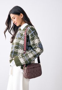 Brun quiltet crossbody-taske med to lynlåslommer, suppleret af en mønstret grøn og cremefarvet sweater og en hvid skjorte under.