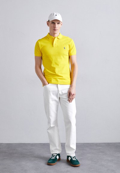 Geltonas polo marškinėliai, balta kepurė, baltos džinsinės kelnės ir žalios nubuko bateliai. Paprastas dizainas su mažu logotipu ant polo ir kepurės.
