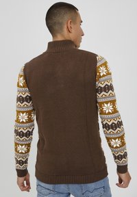 Brązowy sweter wykonany na drutach z wysokim kołnierzem, z wzorzystymi żółtymi, białymi i brązowymi rękawami z motywem płatków śniegu.