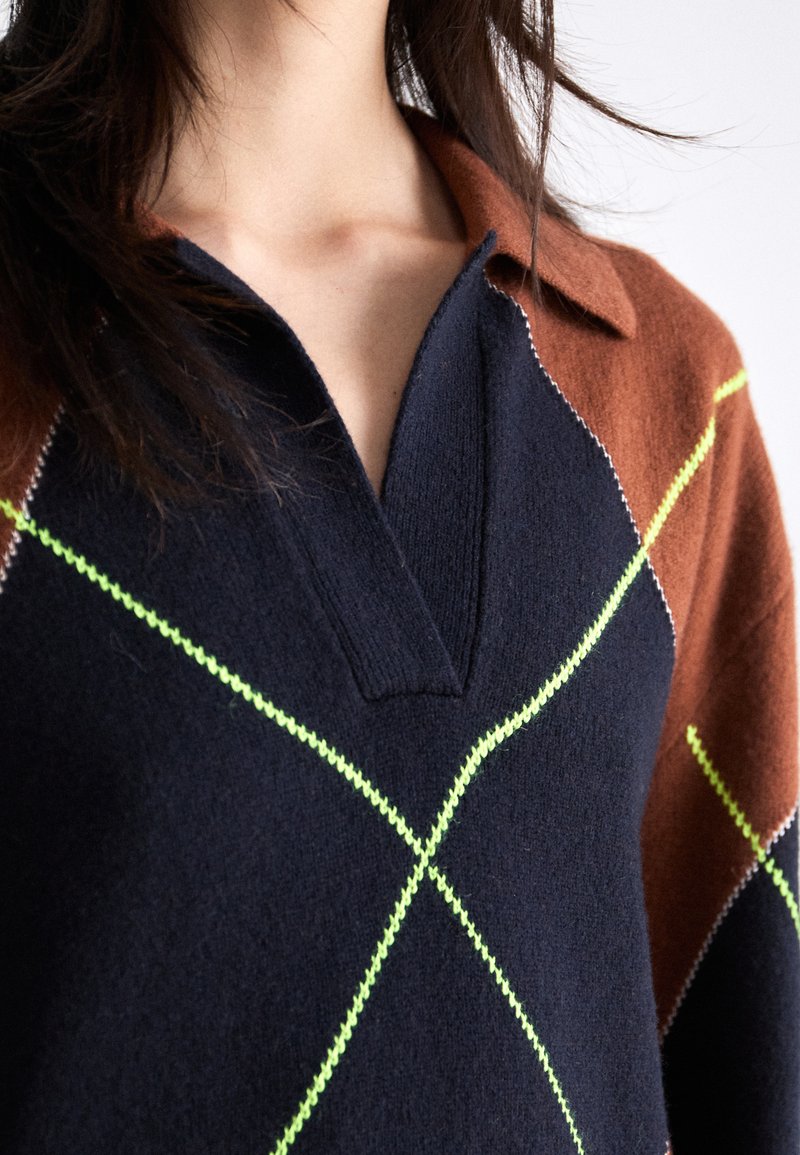 Pull en tricot avec une base bleu marine, des manches marron, un motif en losanges vert et blanc, et un col avec une découpe en V.