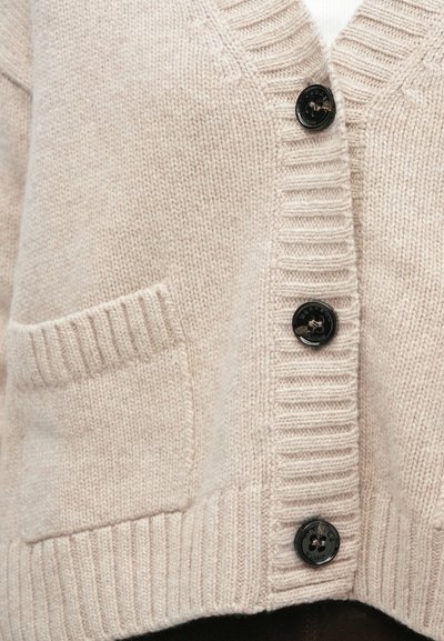 Cardigan de malha bege claro com punhos canelados, decote em V, botões pretos e bolso frontal. Textura aparenta ser macia e quente.