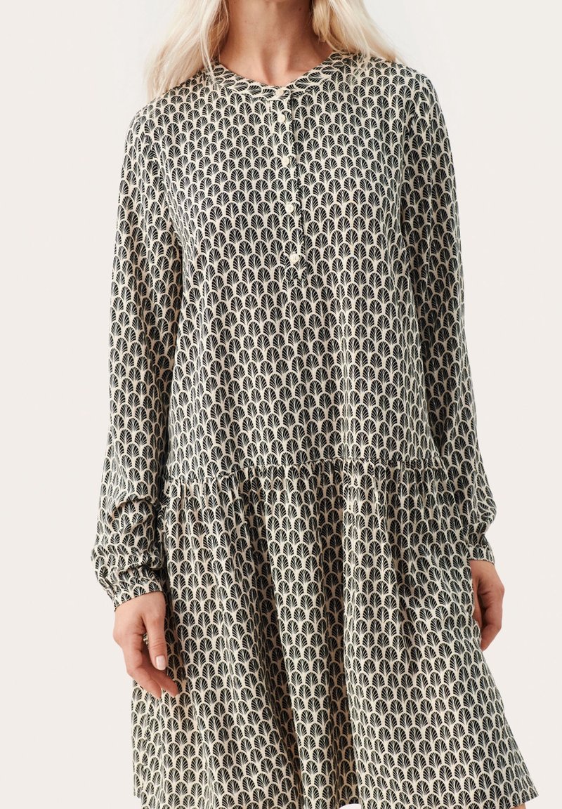 Femme portant une robe à manches longues avec un motif répétitif en forme d'éventail noir et blanc et un col rond boutonné.