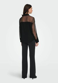 Crna prozirna bluzica s čvrstom pozadinom, dugim rukavima i dugmadi na manžetama. Uparena s crnim hlačama s bijelim točkicama.