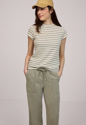 Jeune femme portant un haut à manches courtes rayé beige, un pantalon à cordon de serrage vert olive et une casquette jaune, debout les mains dans les poches.