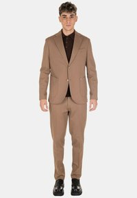 Giovane uomo in piedi, con indosso un completo marrone chiaro abbinato a pantaloni dello stesso colore, scarpe nere e una camicia button down marrone scuro sotto.