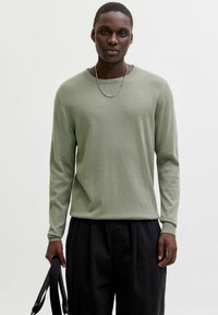 Pull à manches longues vert clair en matière douce, avec un col rond et des poignets côtelés, associé à un pantalon noir, tenu par une main portant une sangle.