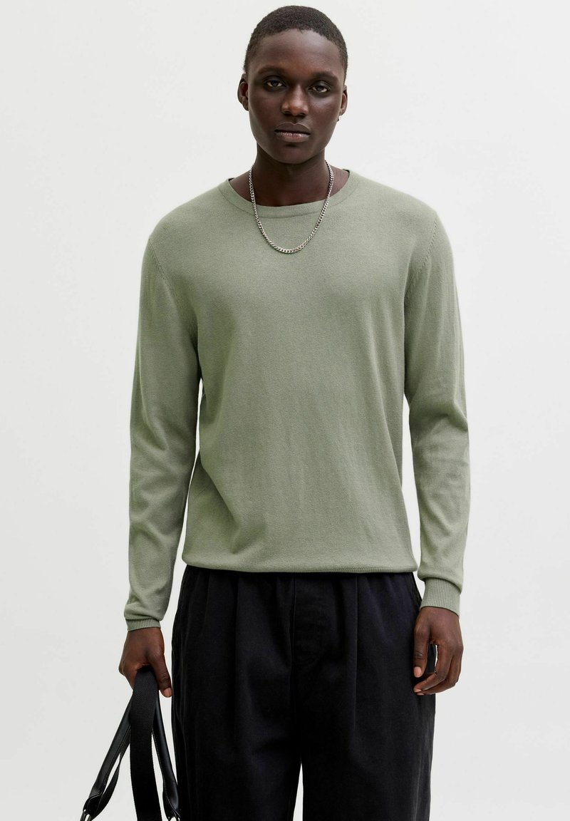 Pull à manches longues vert clair en matière douce, avec un col rond et des poignets côtelés, associé à un pantalon noir, tenu par une main portant une sangle.