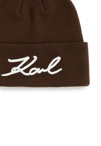 Brun strikket lue med brett som har hvit broderte kursivtekst "Karl" på fronten.