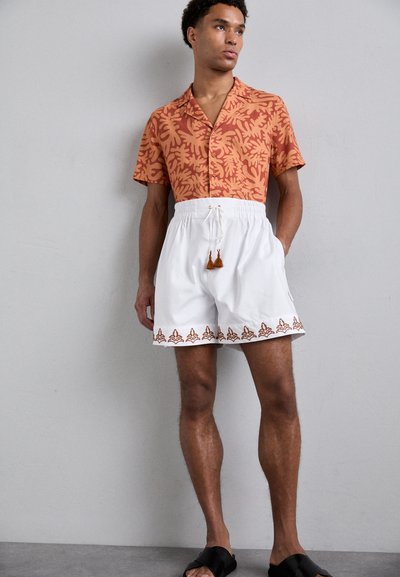 Short-sleeve shirt in orange with leaf pattern, paired with white drawstring shorts featuring brown embroidered detail and tassel accents.