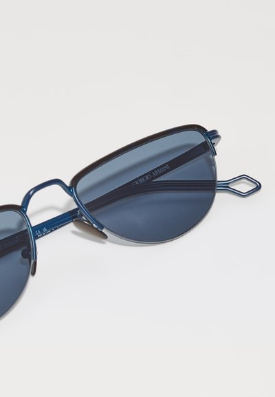 Ochelari de soare tip cat-eye cu nuanțe de albastru au un cadru închis, accente metalice subțiri și un design unic al templelor geometrice. Numele mărcii este vizibil pe lentilă.