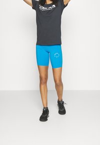 Svart kortärmad t-shirt med grafisk text, kombinerad med ljusblå cykelshorts med en cirkulär logotyp, bärs med svarta sportskor.