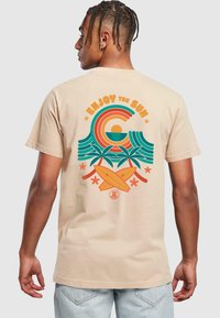 Beige bomulds t-shirt med et farverigt grafisk design af bølger, sol, palmetræer og surfbrætter. Designet inkluderer teksten "Nyd solen".