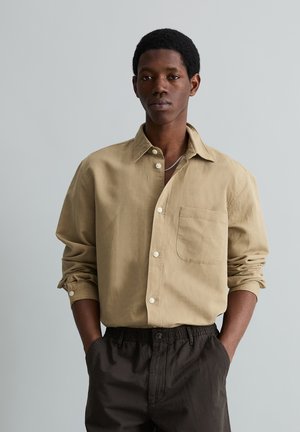 Junger Mann mit kurzen lockigen Haaren, der ein beiges Hemd zum Knöpfen und dunkle Hosen trägt, mit den Händen in den Taschen vor einem grauen Hintergrund stehend.