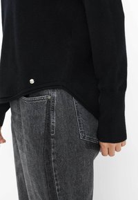 Schwarzer Strickpullover mit langen Ärmeln, der einen Seitenschlitz und ein silbernes Akzentdetail aufweist, kombiniert mit high-waisted grauen Jeans.