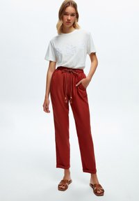 Camiseta blanca con bordado floral, combinada con pantalones rojos con cordón, que cuentan con bolsillos y puños arremangados. Modelo lleva sandalias marrones.