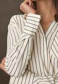 Gestreiftes Blusenshirt in Creme und Schwarz mit V-Ausschnitt, langen Ärmeln, geknöpften cuffs und glattem Stoff, der vertikale Linien und minimale Details zeigt.
