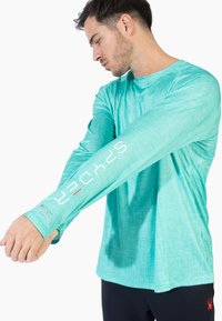 Lys turkis langærmet skjorte lavet af tekstureret stof, med hvide og røde logo detaljer på ærmet samt en rund halsudskæring.