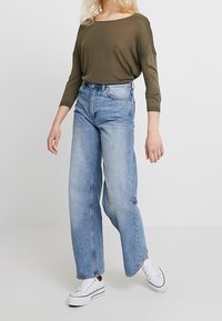 Graugrüner, oversized Pullover mit weitem Ausschnitt, kombiniert mit hellblauen, hochgerafften, locker sitzenden Jeans und weißen Sneakern.