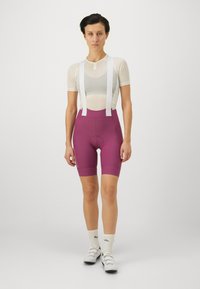 Korte sykkelbukser i dyp lilla stoff, med hvite skulderstropper og polstret sete. Mesh-skjorte under med semi-transparent design.