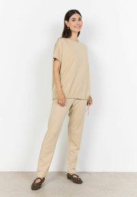 Béžové oversized tričko s krátkými rukávy a stahovací šňůrkou na lemu, v kombinaci s odpovídajícími zúženými kalhotami a hnědými slip-on botami s hroty.