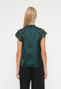 Femme blonde portant un top en satin vert foncé avec des manches courtes en dentelle, vue de dos, associée à un pantalon noir.