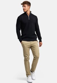 Svart, texturerad tröja med ett vågmönster och halvdragkedja. I kombination med beige byxor och vita sneakers, stående framför en neutral bakgrund.