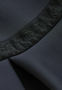 Schwarzer Stoff mit glatter Textur und einem dekorativen schwarzen Netzgürtel mit geprägter Schrift. Das Design konzentriert sich auf subtile Abschlussdetails.